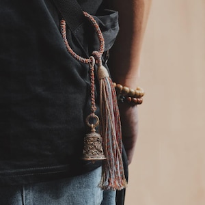 May include: A bronze-colored bell with intricate carvings, suspended by a multi-colored cord and tassel. The bell is attached to a black strap, with a person's arm and a wooden beaded bracelet visible in the background. The bell has a vintage aesthetic.