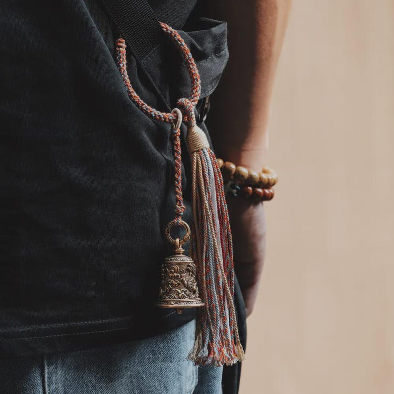 May include: A bronze-colored bell with intricate carvings, suspended by a multi-colored cord and tassel. The bell is attached to a black strap, with a person's arm and a wooden beaded bracelet visible in the background. The bell has a vintage aesthetic.