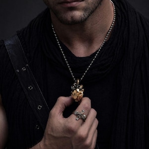 May include: A gold-toned heart-shaped pendant with intricate mechanical detailing suspended from a silver-toned ball chain necklace, paired with a silver-toned ring featuring a detailed cross design with ornate metalwork and small embedded stones.