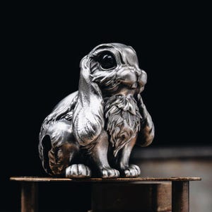 May include: A detailed silver-colored rabbit figurine with large, expressive eyes and long, drooping ears. The sculpture has intricate detailing on its fur and is posed in a seated position. The rabbit is displayed on a wooden surface against a dark background.
