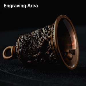 May include: Bronze-colored bell with a dragon design. The bell has a loop for hanging and the text "Engraving Area" at the top. The bell is made of metal and has intricate details.