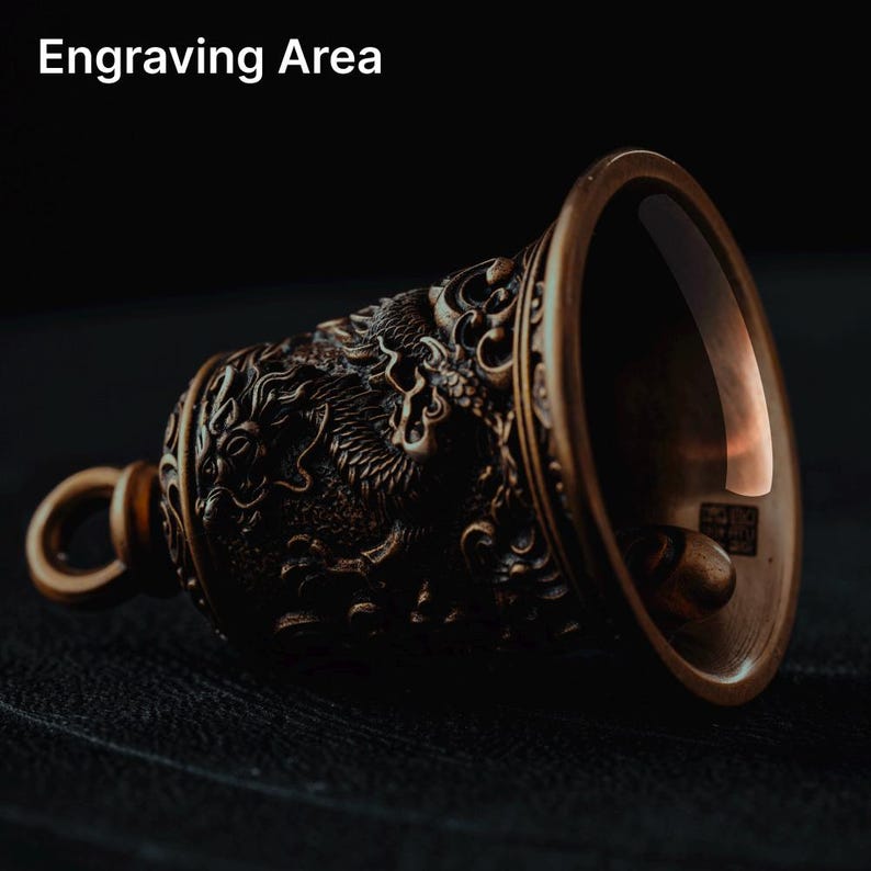 May include: Bronze-colored bell with a dragon design. The bell has a loop for hanging and the text "Engraving Area" at the top. The bell is made of metal and has intricate details.