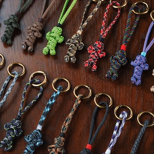 May include: A collection of keychains with braided cord lanyards and metal rings. The keychains feature various colors, including green, brown, red, blue, and black. The braided cords are knotted into small figures.