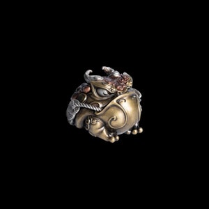 Könnte beinhalten: Eine goldene, silberne und kupferne Froschfigur mit filigranen Details. Der Frosch sitzt auf seinen Hinterbeinen, mit offenem Maul und geschlossenen Augen. Er hat ein Seil um den Hals und eine Münze im Maul.