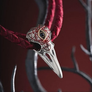 May include: A silver-toned bird skull pendant featuring intricate red etched line patterns and a central gold-toned eye motif with a dark stone inlay. The pendant is suspended from a thick, braided deep red cord necklace.