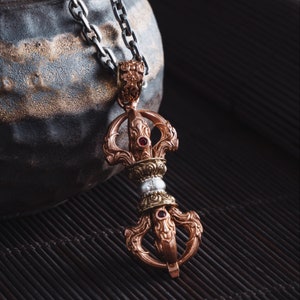 以下が含まれることがあります： 複雑なディテールが施された銀と銅のペンダント。赤い宝石と回転する車輪のデザインが特徴です。ペンダントは銀のチェーンからぶら下がっています。