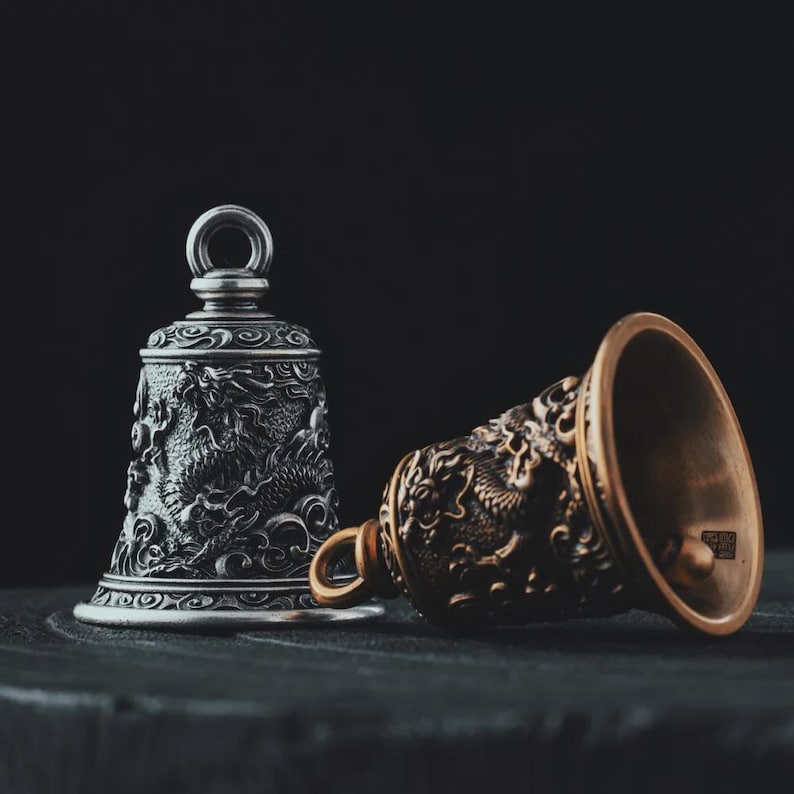 May include: Two decorative metal bells with dragon motifs. One bell is silver and upright, while the other is bronze and lying on its side. Both bells have intricate carvings and a dark background.