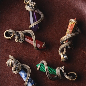 May include: Five gemstone pendants with bronze snake detailing. The pendants feature various coloured gemstones, including purple, red, and green. The jewellery is displayed on a brown leather surface, showcasing the intricate design.