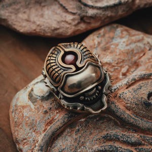 May include: A detailed, steampunk-style ring with a central red gemstone. The ring features intricate metalwork in bronze and silver tones, with a textured, mechanical design. The ring is set against a natural stone background.