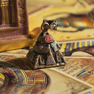 May include: A bronze-colored pyramid-shaped decorative object with hieroglyphs and colorful accents. The pyramid has a circular emblem with the number 13 in red. The object is on a surface with ancient Egyptian-style artwork.