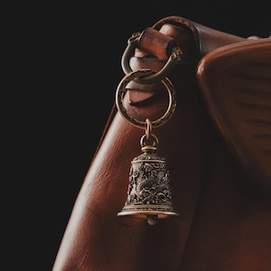 May include: A detailed close-up of a brass bell keychain with intricate dragon and floral designs. The bell is suspended from a ring attached to a leather strap. The background is a dark, blurred leather surface.