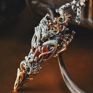 Könnte beinhalten: Aufwendiger silberner Anhänger mit goldenen Akzenten und einem roten Mittelteil. Der Anhänger weist aufwendige Filigranarbeiten und einen kleinen grünen Edelstein auf. Das Design umfasst einen stilisierten Tierkopf und florale Motive.