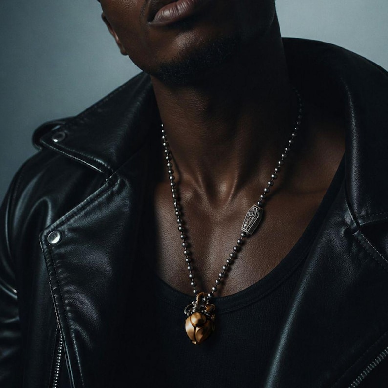 May include: A silver ball chain necklace featuring a small, rectangular coffin-shaped charm and a larger, metallic gold-toned anatomical heart pendant. The necklace is worn over a black tank top and a black leather jacket with silver metal snaps.