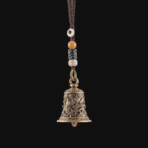 May include: Bronze-colored bell with intricate dragon carvings, suspended by a brown braided cord and decorative beads. The bell has a flared base and a small clapper. The beads are in shades of white, brown, and black.