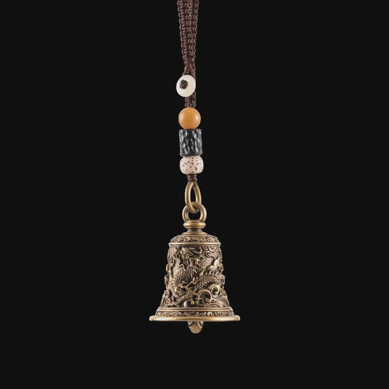 May include: Bronze-colored bell with intricate dragon carvings, suspended by a brown braided cord and decorative beads. The bell has a flared base and a small clapper. The beads are in shades of white, brown, and black.