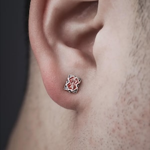 Può includere: Un piccolo orecchino a bottone quadrato in argento con un design rosso e bianco. L'orecchino è indossato su un orecchio. Il design presenta un elemento centrale rosso con accenti bianchi e neri. L'orecchino ha un motivo geometrico dettagliato.