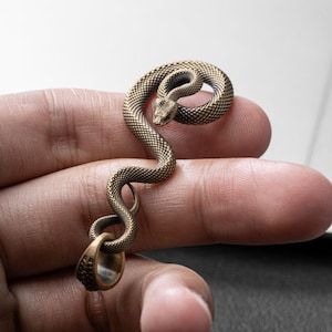 May include: A bronze-toned metal pendant shaped like a coiled snake with a textured, scale-like surface. The snake's body curves into an S-shape, ending in a smooth, circular loop at the bottom designed for attachment to a chain or cord.