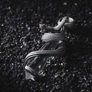 May include: A silver-toned metal pendant featuring a detailed snake coiled around a clear, elongated, hexagonal crystal point. The snake has visible scales and an open mouth, with the body wrapping multiple times around the transparent stone.
