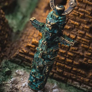 May include: A vertical pendant featuring a weathered teal and bronze finish with intricate relief carvings of mythical creatures and geometric patterns. The cylindrical object includes a circular metal loop at the top for attachment.