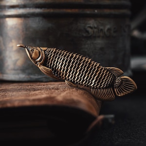 May include: A detailed bronze-coloured fish sculpture with textured scales and fins. The fish is positioned on a wooden surface, with a blurred, dark background. The sculpture has an antique or vintage aesthetic.