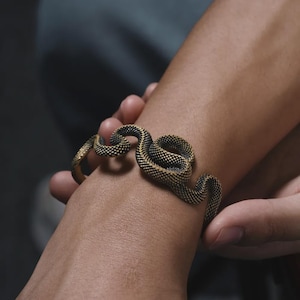 May include: A detailed bronze-colored snake bracelet. The bracelet wraps around the wrist, featuring a textured, scale-like pattern in black and gold. The snake design is a unique piece of jewelry.