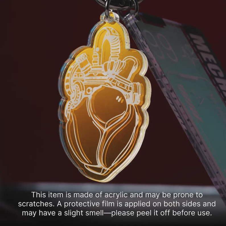 May include: A translucent amber-colored acrylic keychain featuring a white line-art illustration of a mechanical heart. The design includes intricate pipes and valves, suspended from a metal ring. The item has a protective film on both sides.