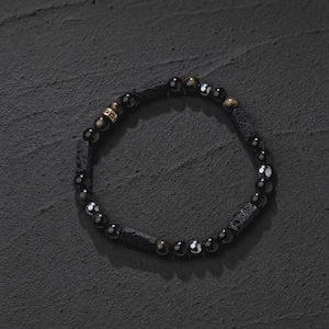 May include: A stretch bracelet featuring alternating black lava rock beads and smooth, polished black obsidian beads. The design includes a single small, cylindrical gold-toned metal accent bead with engraved detailing, creating a contrast in texture and colour.