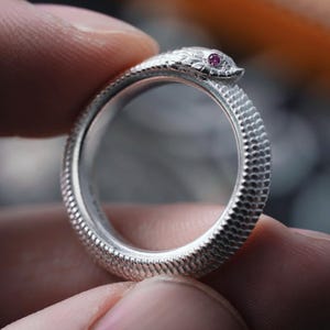 May include: A silver ring shaped like a snake, featuring a textured body and a small, round, pink gemstone set in the snake's head. The ring is held between two fingers.