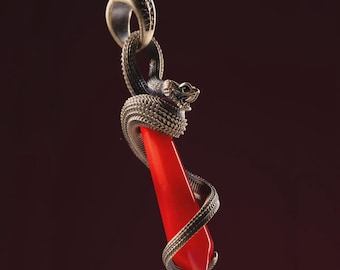 May include: A silver-toned pendant featuring a red, elongated gemstone embraced by a detailed snake. The snake's body coils around the stone, with its head positioned at the top. The pendant has a loop for a chain.