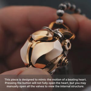 May include: A metallic pendant featuring a gold-toned, multi-faceted exterior with mechanical gear-like details. The object is suspended from a beaded chain. Text on the image reads: This piece is designed to mimic the motion of a beating heart.