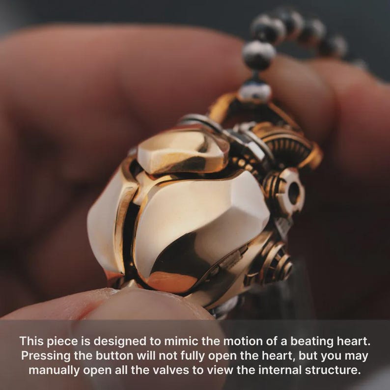 May include: A metallic pendant featuring a gold-toned, multi-faceted exterior with mechanical gear-like details. The object is suspended from a beaded chain. Text on the image reads: This piece is designed to mimic the motion of a beating heart.