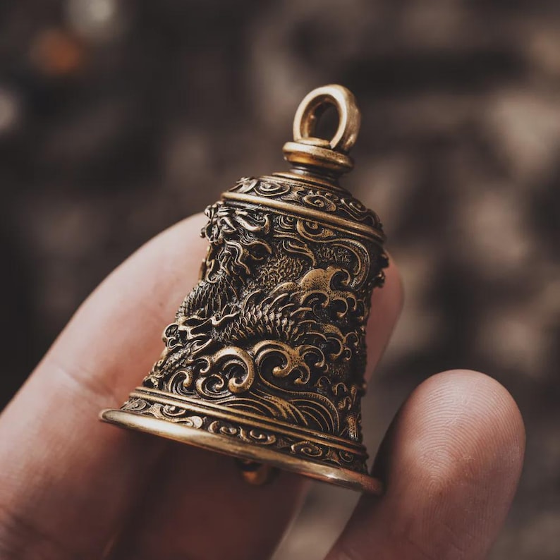 May include: A detailed brass bell with an ornate dragon design. The bell has a loop at the top for hanging and intricate carvings. The bell is held in a hand, showcasing its size and craftsmanship.