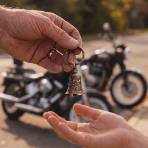 May include: A bronze motorcycle bell with intricate detailing is held in a hand, with another hand reaching out to receive it. The bell is suspended from a ring and features a detailed design. A motorcycle is blurred in the background.