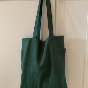 May include: A dark green tote bag made of textured fabric hangs from a dark knob. The bag has long straps and a rectangular shape. A small, rectangular tag is sewn on the side of the bag.