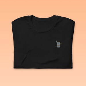 Może przedstawiać: Czarny t-shirt z małym haftem przedstawiającym srebrny dzbanek do kawy z brązowym uchwytem i parą unoszącą się z dzióbka.