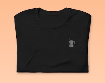 以下が含まれることがあります： 小さな刺繍が施された黒いTシャツ。銀色のコーヒーポットに茶色の取っ手と注ぎ口から出る蒸気が描かれている。