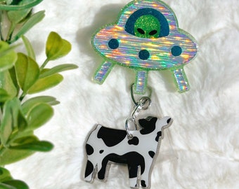 May include: A holographic alien spaceship badge reel with a green alien and a black and white cow charm. The badge reel is attached to a silver-colored metal clip. The background is a white, fluffy surface.