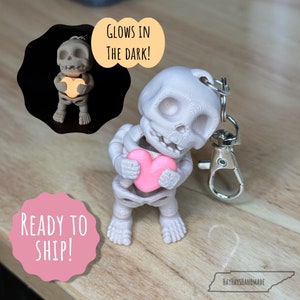 May include: A white glow-in-the-dark skeleton keychain holding a pink heart. The keychain has a silver metal clasp. "Glows in the dark!" and "Ready to ship!" are written on the image.