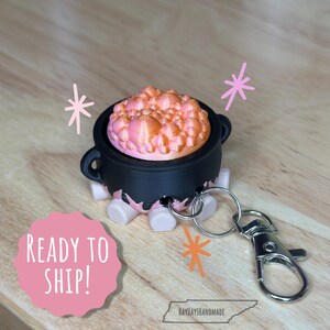 May include: A black cauldron keychain with a pink and orange bubbling potion inside. The cauldron is on a white stand with a silver key chain attached.