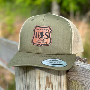 May include: A green and beige trucker hat with a brown leather patch that says "Forest Service U.S. Bigfoot Department".