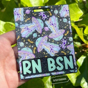 May include: A black and glittery badge holder with a space-themed design featuring UFOs, aliens, planets, and stars. The badge holder has the text "RN BSN" in light green. The bottom of the badge holder has the text "HAYHAYSHANDMADE".