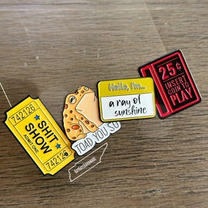 May include: A set of five enamel pins featuring a yellow ticket with the text "742126 SHIT SHOW 742126" and the words "ADMIT ONE", a yellow toad with the text "TOAD YOU SO", a yellow rectangle with the text "Hello, I'm... a ray of sunshine", a red book with the text "25¢ INSERT COIN TO PLAY", and a black and white outline of the state of Tennessee with the text "HAYHAYSHANDMADE".