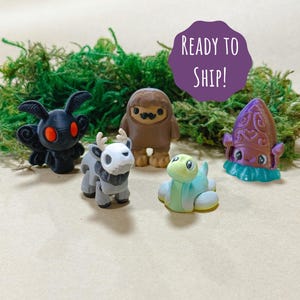 May include: A collection of five miniature figurines, including a black moth with red eyes, a gray deer-like creature, a brown Bigfoot, a light blue turtle, and a purple squid. The figurines are displayed on a bed of green moss.