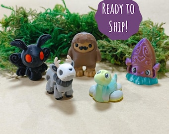 May include: A collection of five miniature figurines, including a black moth with red eyes, a gray deer-like creature, a brown Bigfoot, a light blue turtle, and a purple squid. The figurines are displayed on a bed of green moss.
