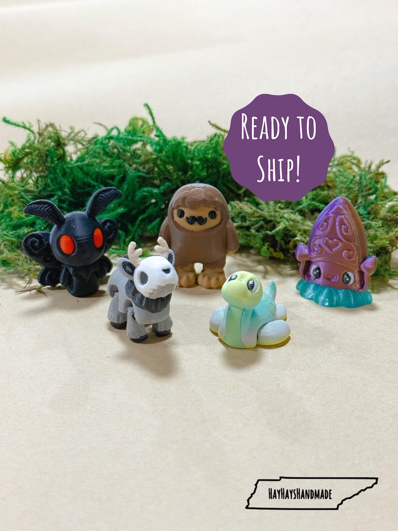 May include: A collection of five miniature figurines, including a black moth with red eyes, a gray deer-like creature, a brown Bigfoot, a light blue turtle, and a purple squid. The figurines are displayed on a bed of green moss.