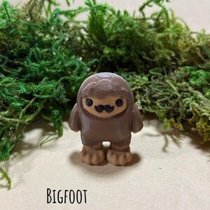 May include: A brown Bigfoot figurine with a tan face and feet, and a black mustache. The word "BIGFOOT" is printed in black below the figurine. The HayHaysHandmade logo is in the bottom right corner.