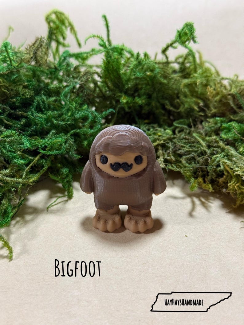 May include: A brown Bigfoot figurine with a tan face and feet, and a black mustache. The word "BIGFOOT" is printed in black below the figurine. The HayHaysHandmade logo is in the bottom right corner.