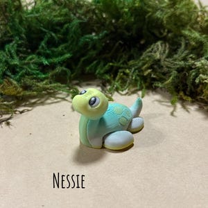 May include: A handmade polymer clay sculpture of a turtle, in shades of blue, green, and yellow. The word "NESSIE" is written in black script below the turtle. The HayHaysHandmade logo is at the bottom.