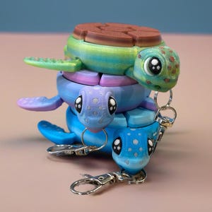 May include: Three stacked turtle keychains. The top turtle is green with brown accents, the middle turtle is purple with blue accents, and the bottom turtle is blue with white accents. Each turtle has a black eye with a white pupil and a silver keychain ring.
