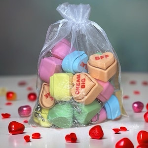 May include: A white mesh bag filled with colorful heart-shaped candies. The candies have different messages on them, including "Dream Big", "BFF", and "Love".